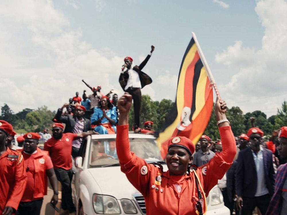 A black man in a suit stands on a ute, parading, his supporters in red caps and surrounding the car