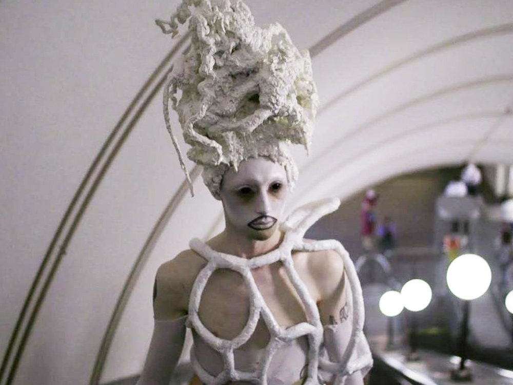 A person with white makeup, black eyes, a head crown that looks like a papier mache beehive, and a white coral-like structure over their chest