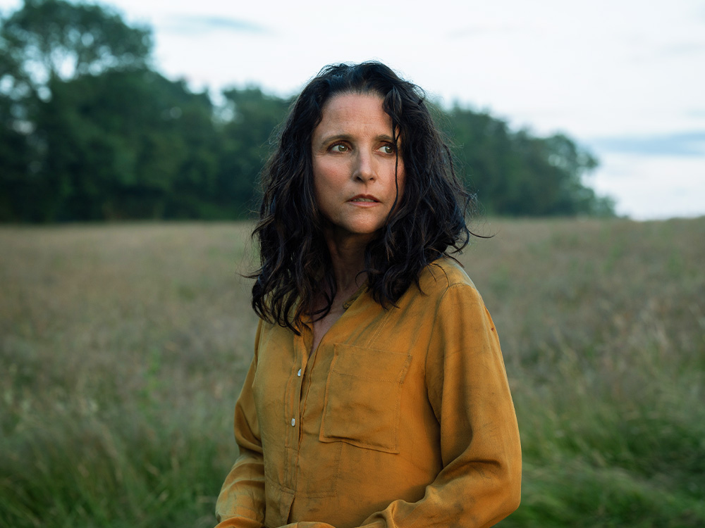 A woman stands in a field of grass, looking off at the sky