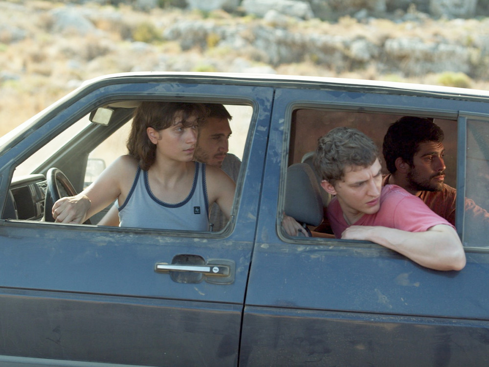 A group of young people in a dusty car. They're looking out at something behind them.