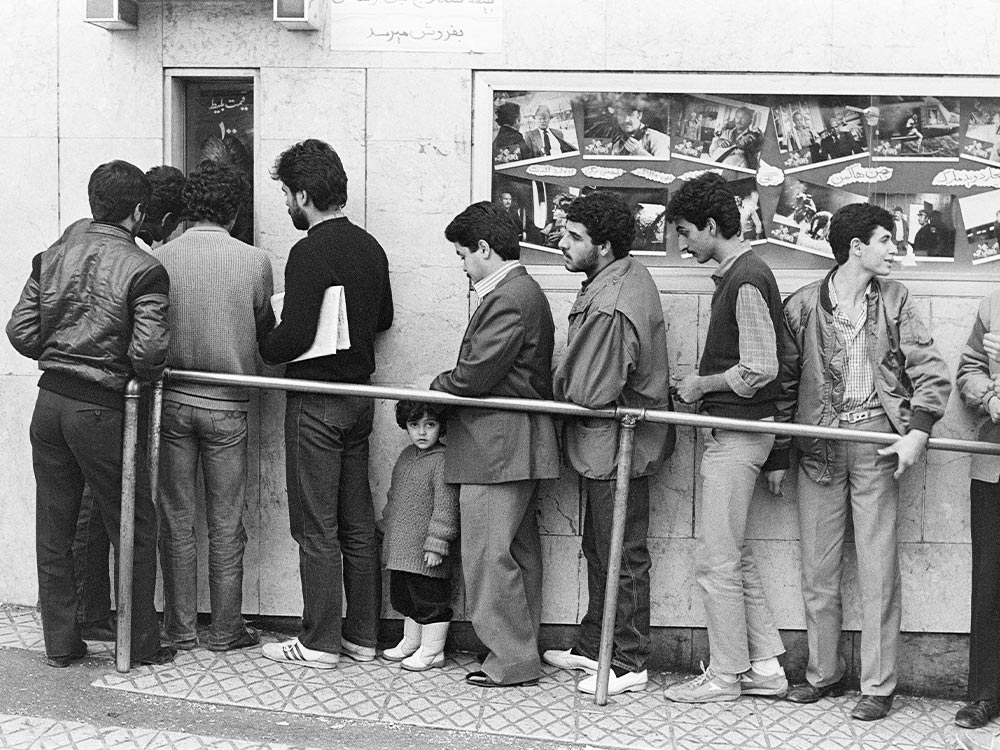 A line of men and a child wait to enter a cinema. Film stills and Arabic text decorate a board outside the building