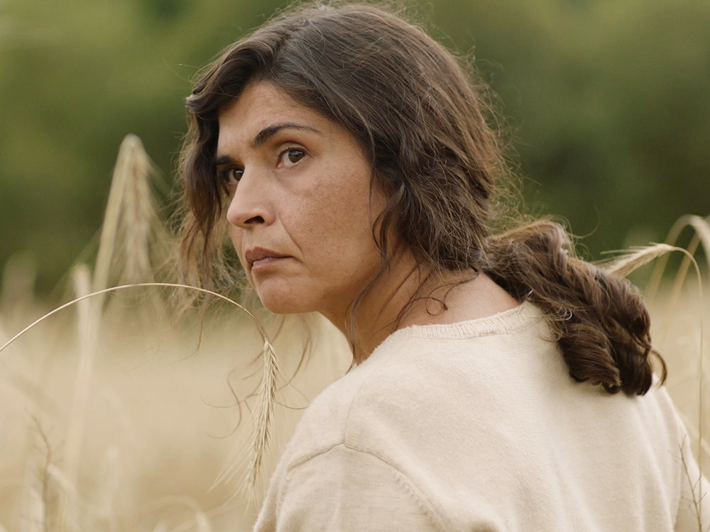 A woman with long hair stands in a wheat field, looking back