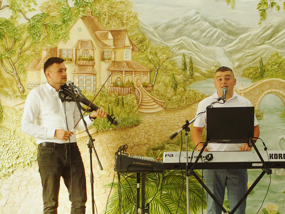 Two men playing muic, one on a black electric violin, the other on a KORG keyboard. Both are singing into mics, against a painted backdrop of a house inside the woods, by a river, a mountain range in the back