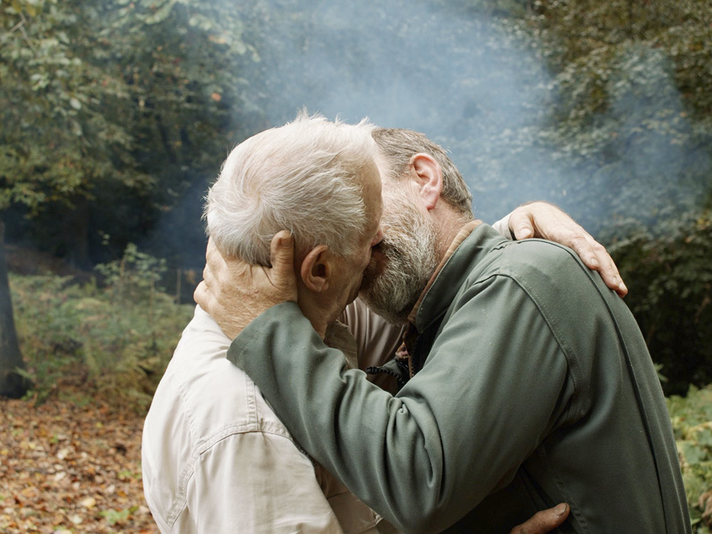 Two men with greying hair kiss passionately in a forest