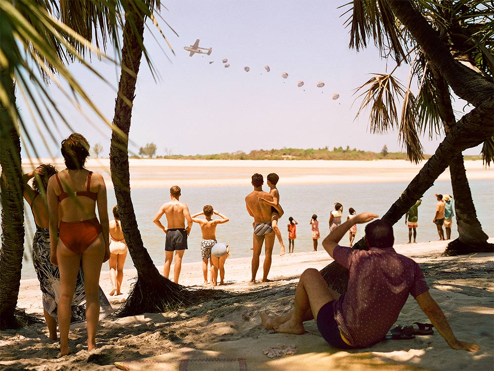 A group of beachgoers watch a plane drop parachute packages across the water