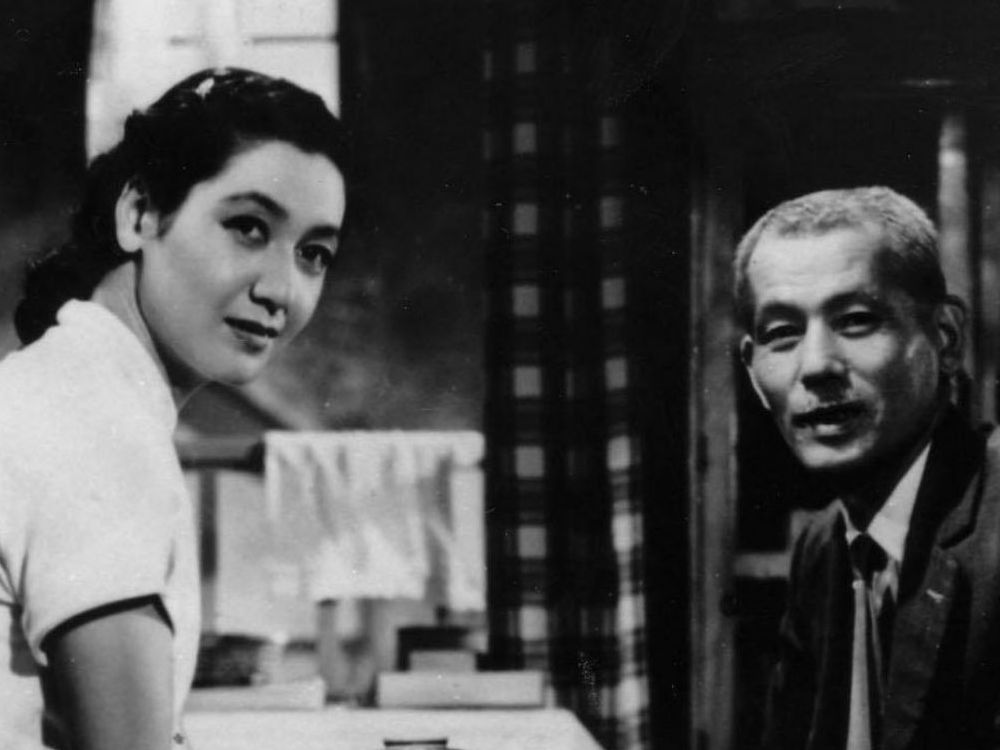 In black and white, a young Japanese woman and an older Japanese man in a suit sit in their home, smiling