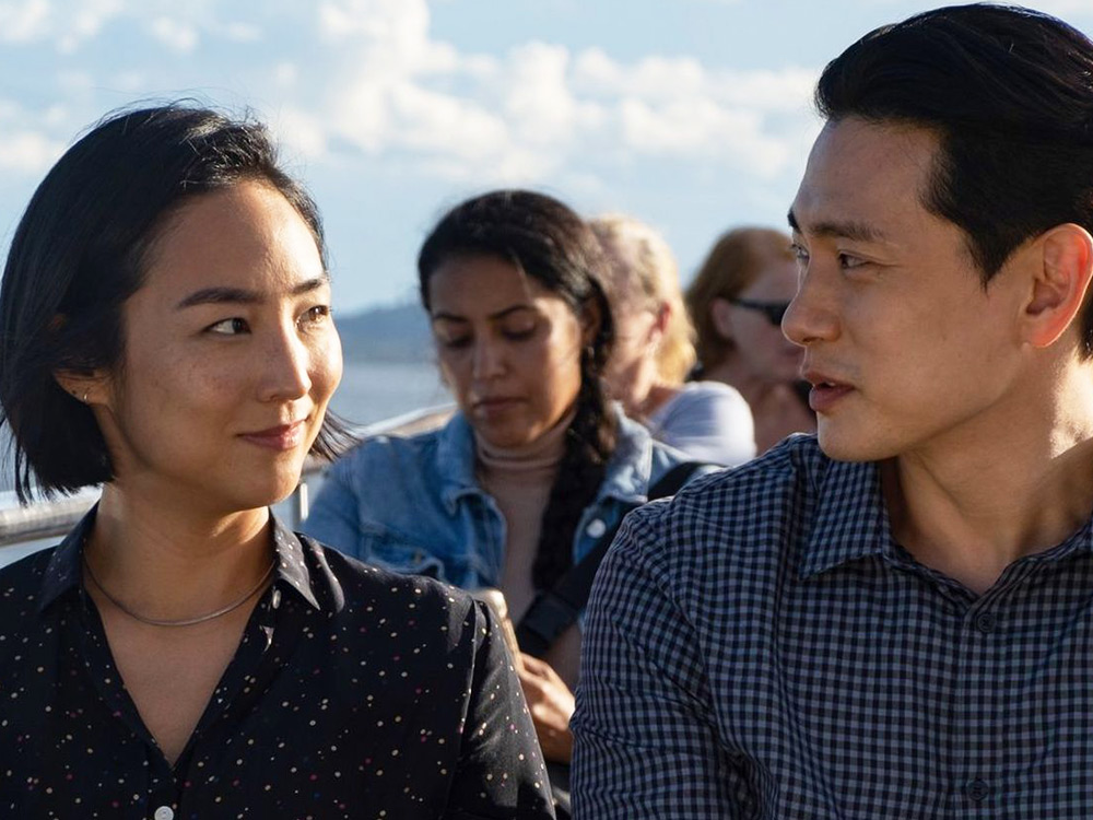 A Korean woman and a Korean man look at each other longingly, as they sail on a boat in the New York City harbour