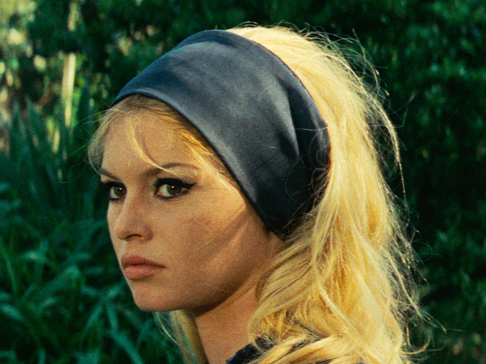 A woman with blonde hair and a black headband stands against green trees and bushes
