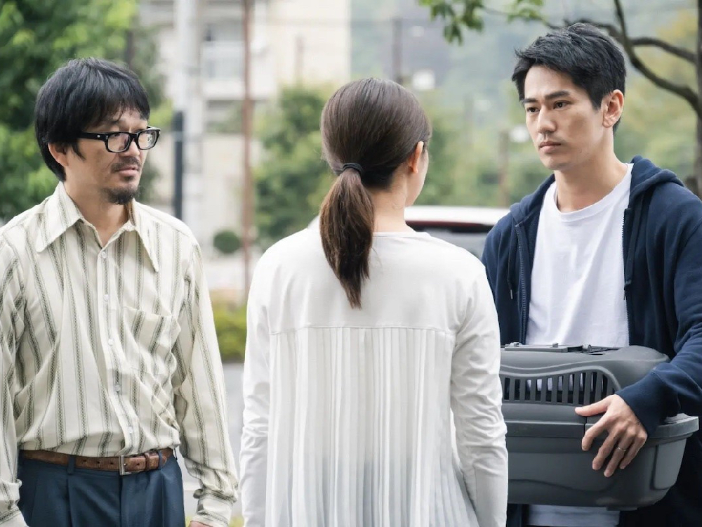 Three people stand outside, mid-conversation. One of them holds a box which could be used to take an animal to the vet.