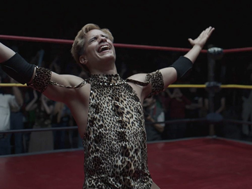 Cassandro kneels in the wrestling ring, arms in the air in elation, dressed in a leopard print sleeveless outfit and elbow pads.