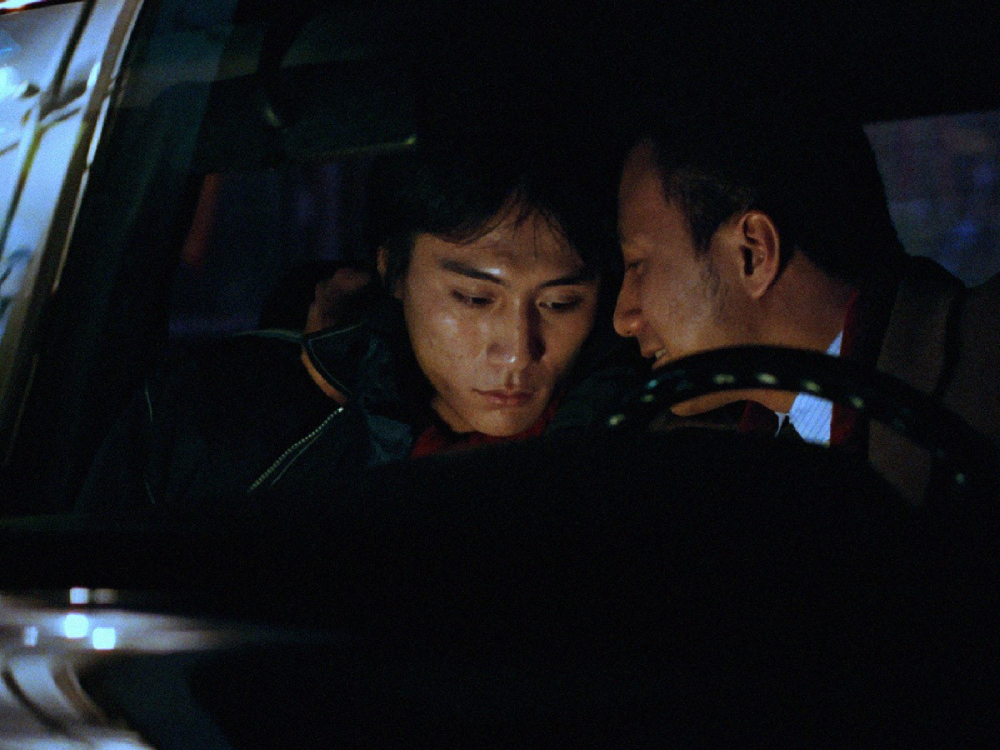 Two men are close to one another in a car, one looks happily at the other. The other man's eyes are low, he doesn't look happy. It is night time.