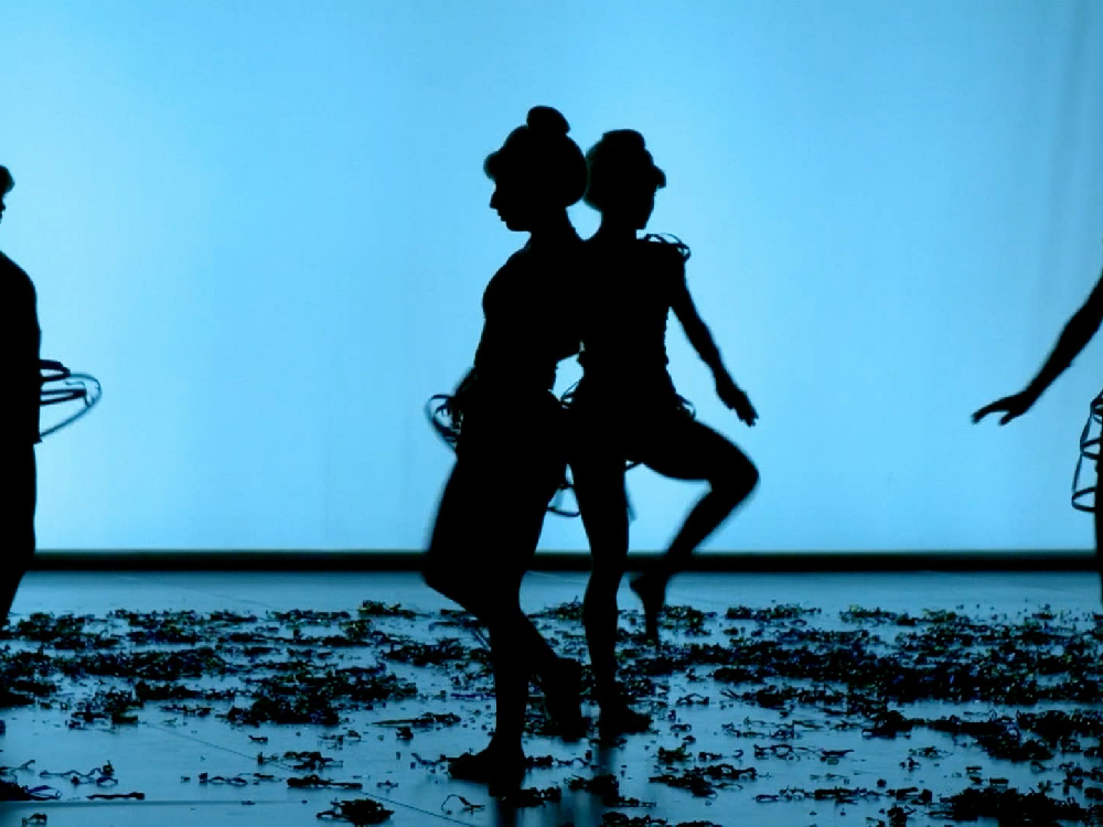 A performance space, something litters the floor, two dancers are silhouetted in movement against a pale blue backdrop