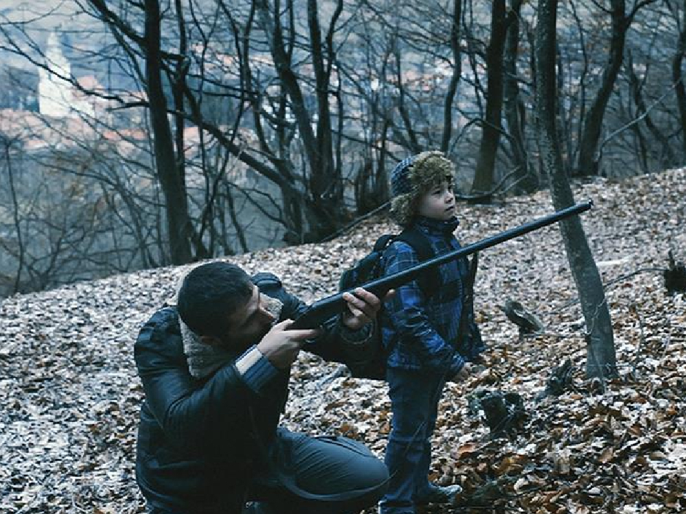 A man crouches on a leaf covered forest floor, pointing a rifle off screen to the right. A small boy stands next to him looking in the same direction.