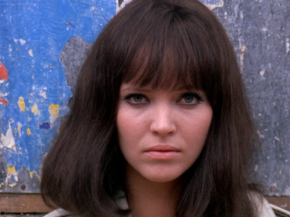 a white woman with dark brown hair, with a fringe, looks directly to camera. The background is a wall that remnants of posters and paint.