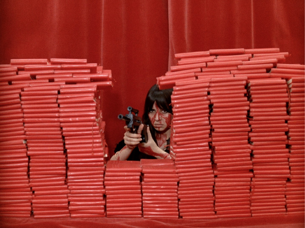 A white woman points a gun out from between stacks of red books, her face and arms have blood on, the background is a red curtain