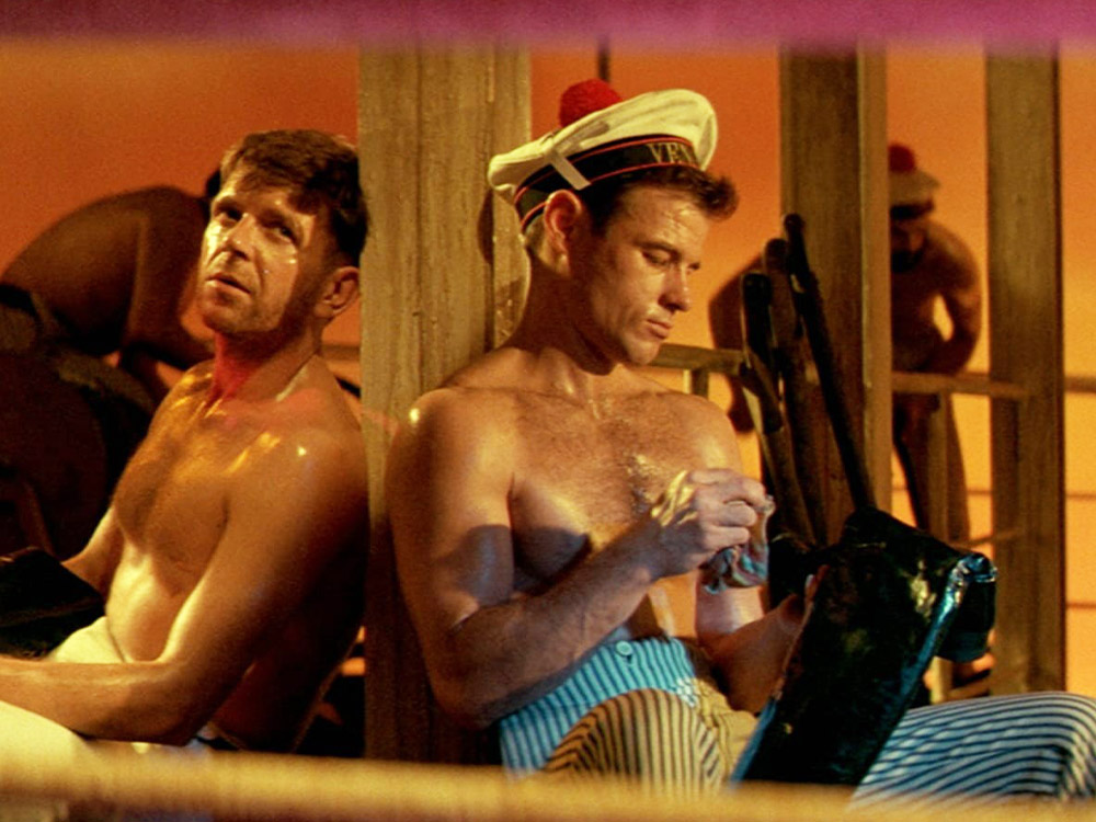 Two white men sit leaning against a wooden beam. One wears a sailor hat and is polishing a boot. They are both topless and sweating.