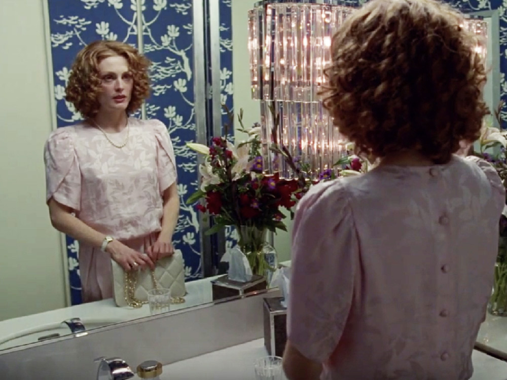 A white woman with curled hair looks at herself intensely in a bathroom mirror, she holds a small handbag