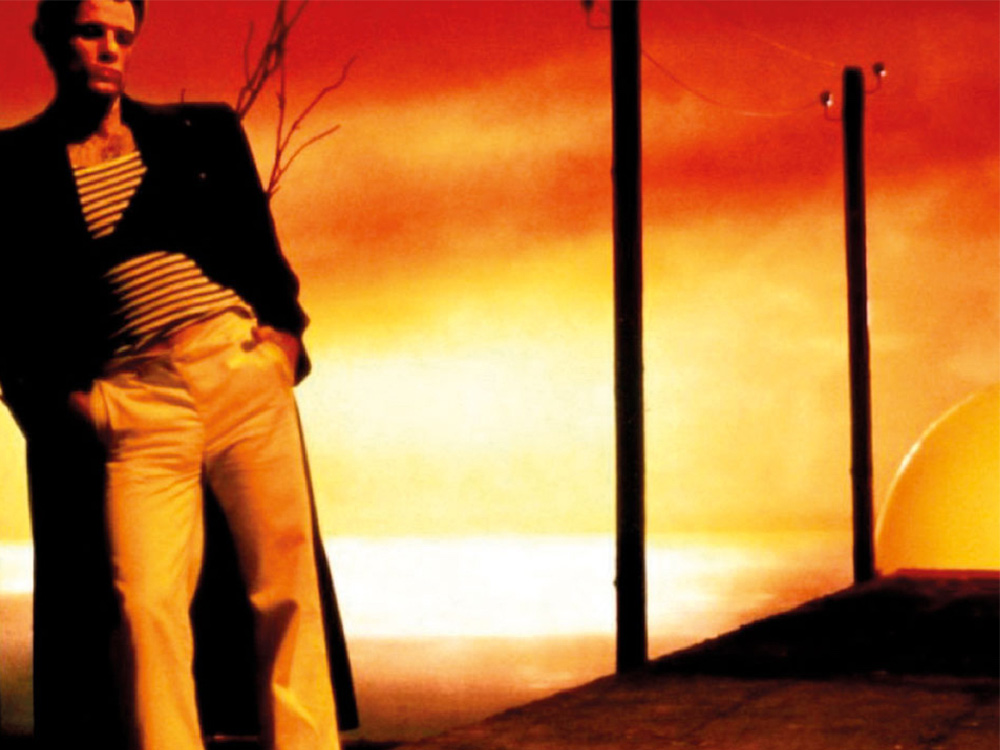 a white man leans thoughtfully against a post. The background is red, orange, yellow - similar to a sunset