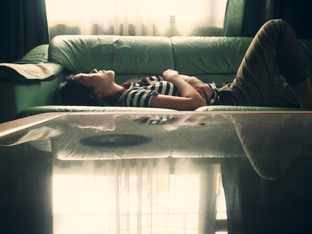 A woman lies down on a couch in front of a window, light reflecting off the in front of her