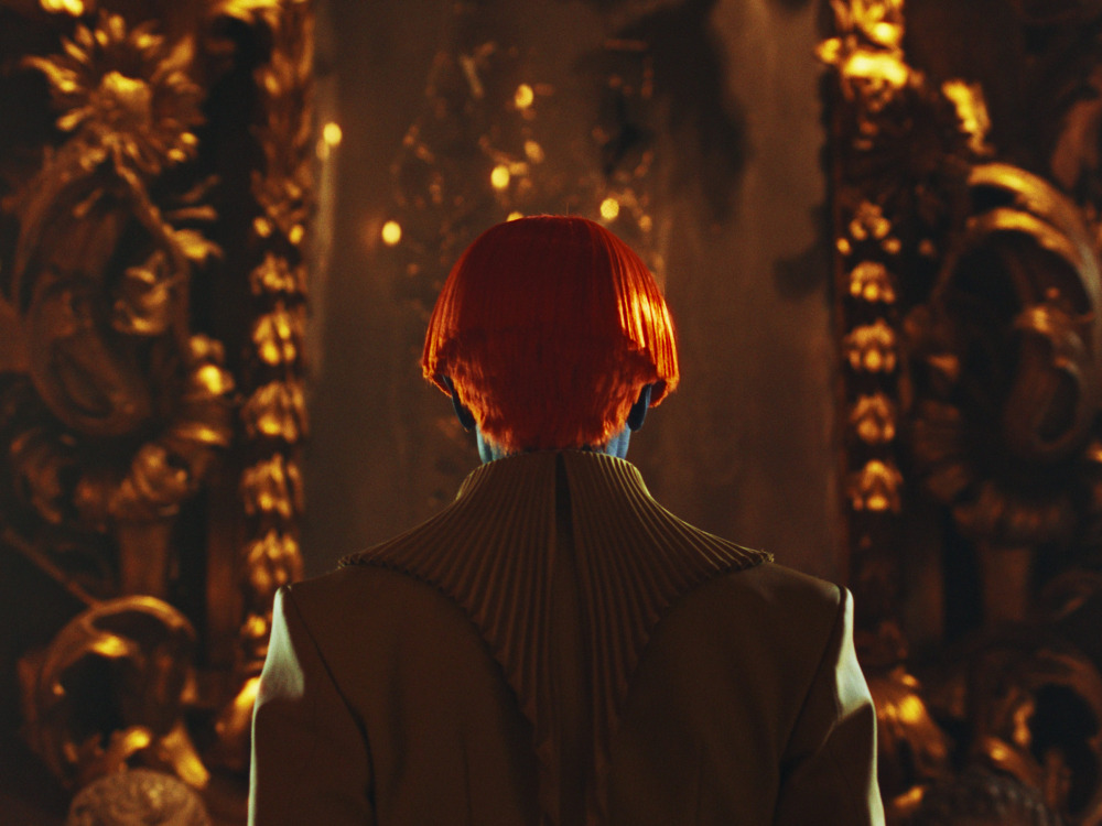 The back of a person's head, their hair is orange with a bowl cut, skin blue. They're wearing a white sci-fi regal suit and staring into a space between two ornate gold columns