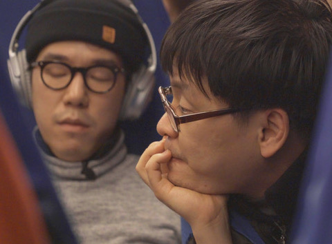 A Korean man with glasses concentrates, his hand on chin, while another sits in the back, closing his eyes and listening quietly to his headphones