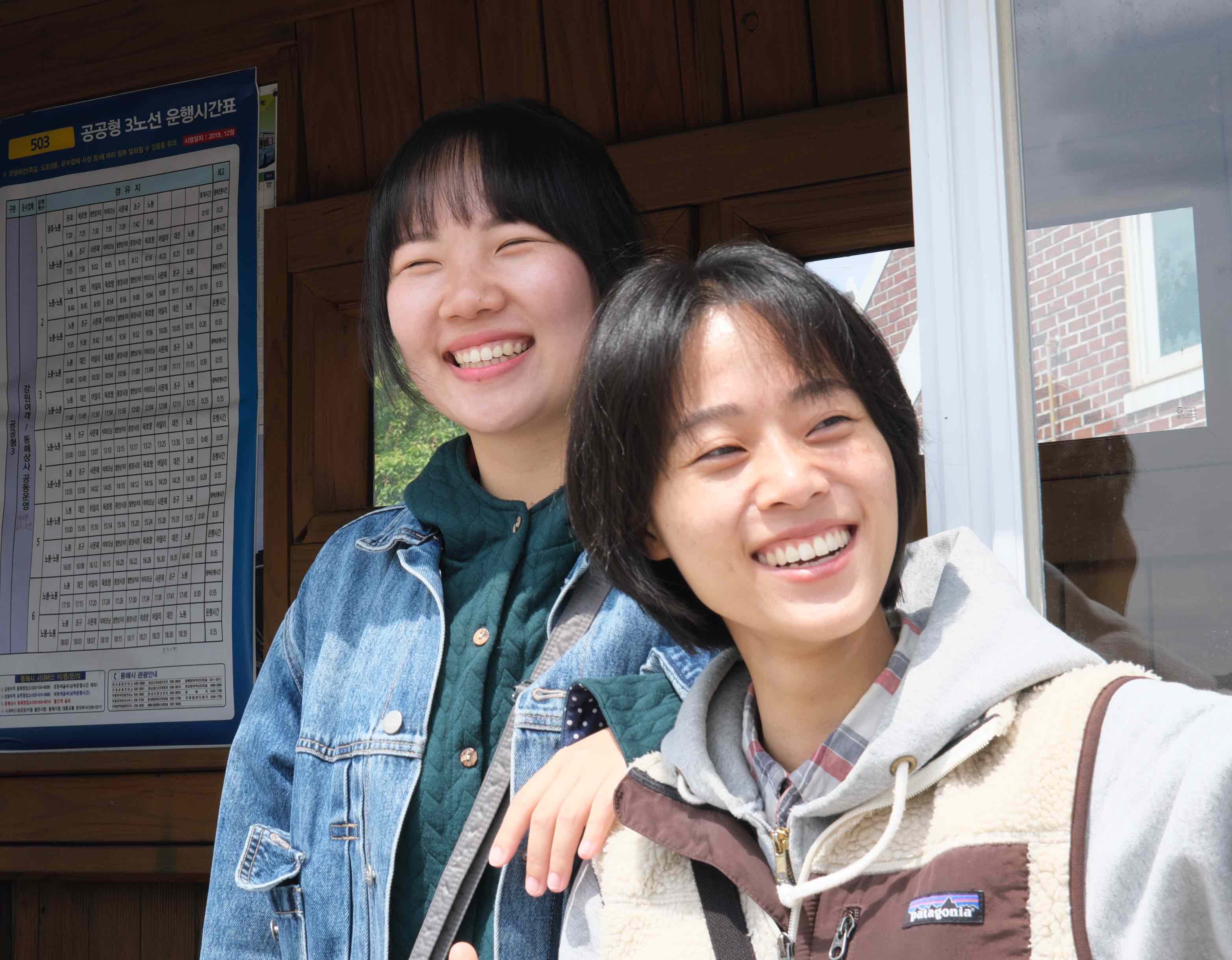 Two young smiling Korean people stand by a wooden shack