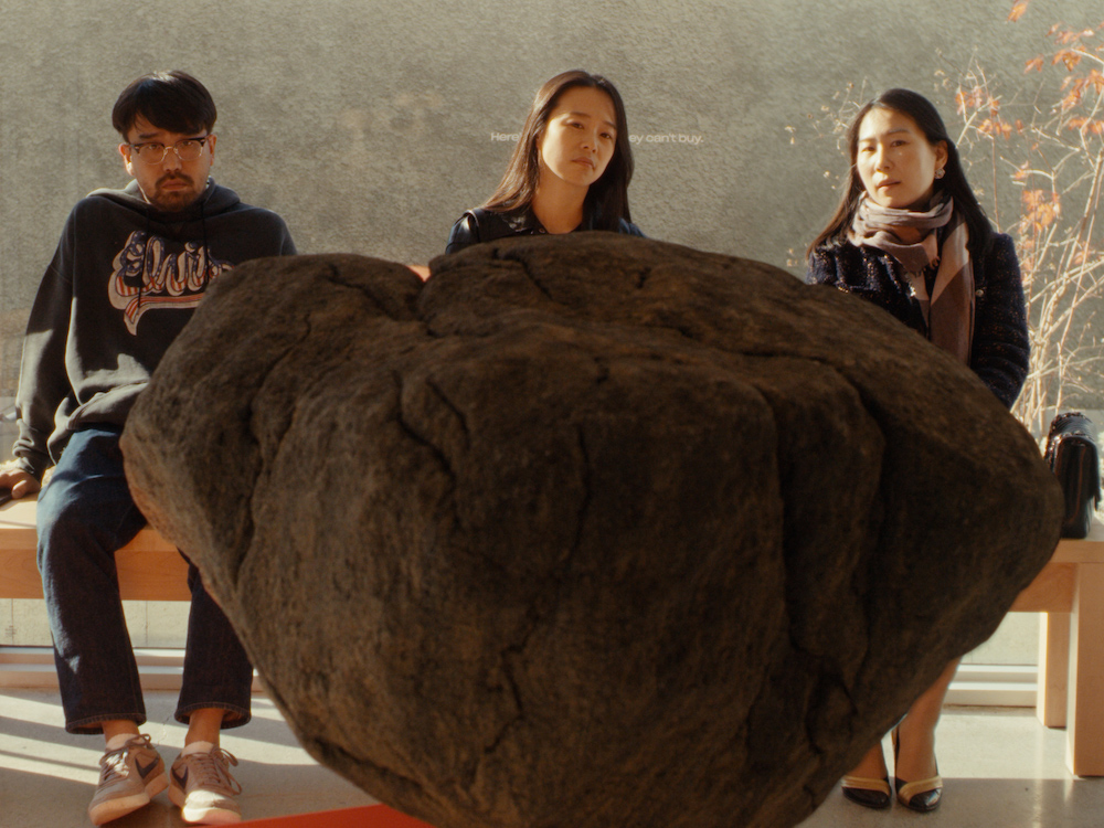 Three Korean people look confused. They sit on a bench behind a huge rock statue