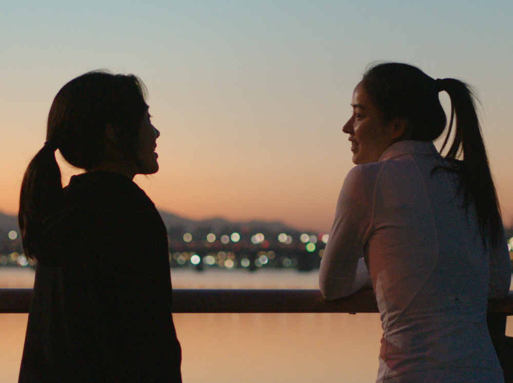 Two Korean women in running gear stand on the bridge, talking as the sun sets to orange and pink