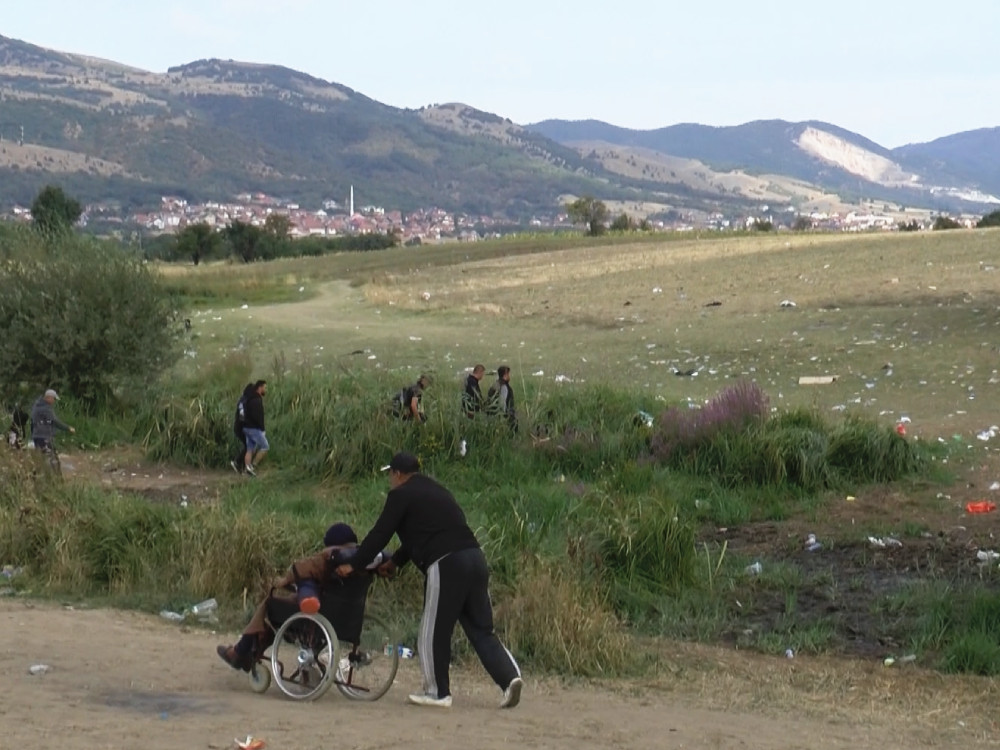 A man wheels an elderly woman on a wheelchair next to a grass field and hills