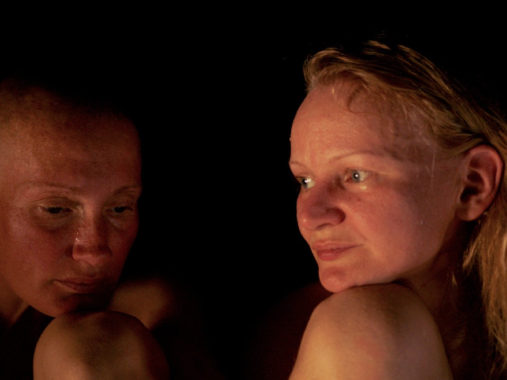 two white female presenting people sit in the darkness of a sauna, one looks down and the other looks off screen.