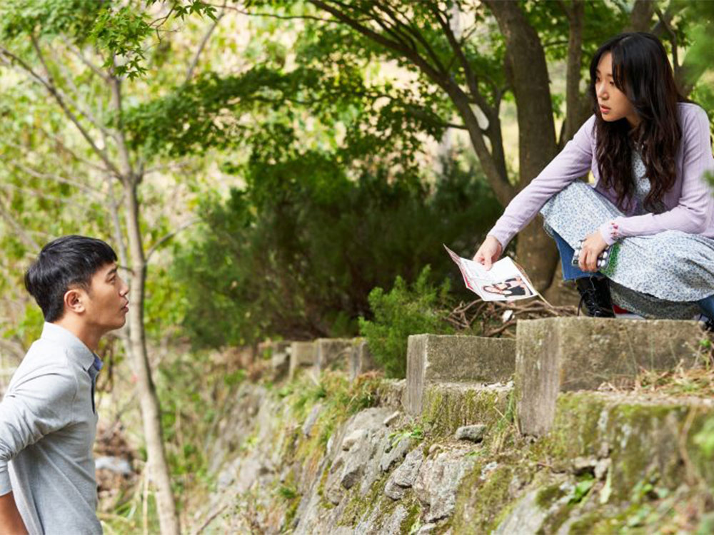 A Korean woman kneels on top of a forest path, holding a photo, showing it to a man below