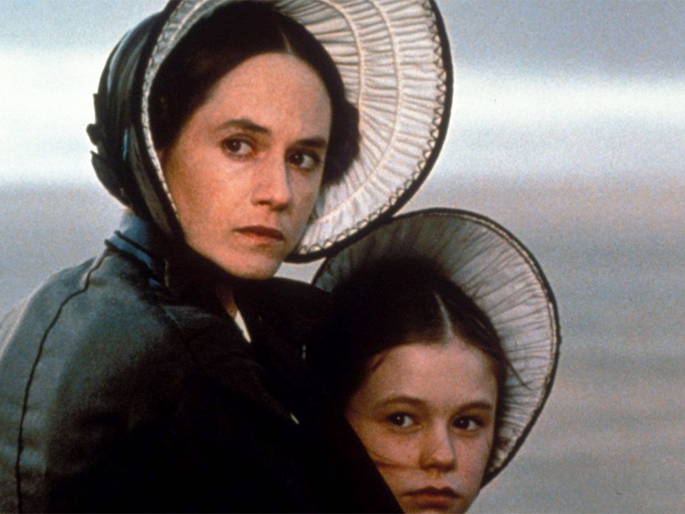 A young white woman and her daughter wear matching bonnets. They stand against a murky background