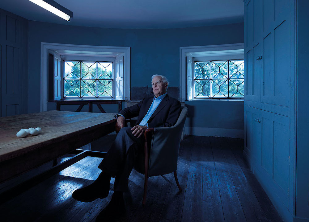 A man with white hair in a suit sits in a large wooden living room