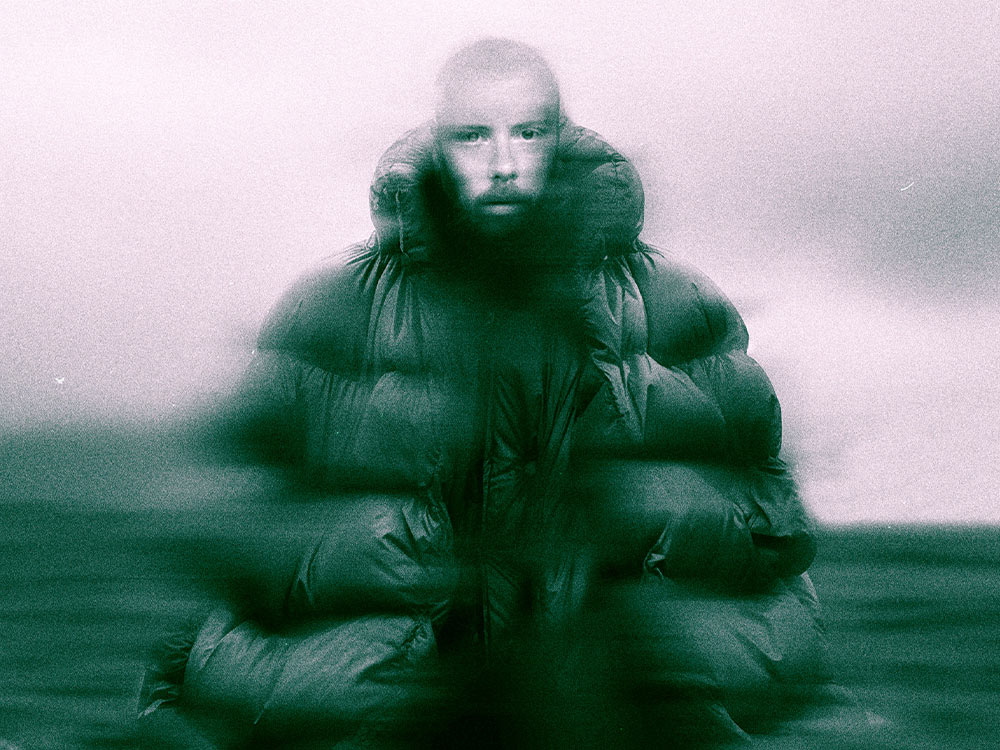 Press pic of Forest Swords. He is bald, white, standing with a large puffer jacket against a dull grey and green blurred background