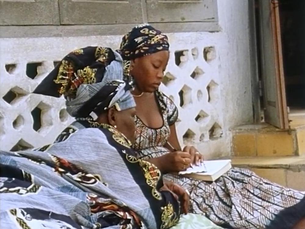 Two African women write on paper, sitting outside their home