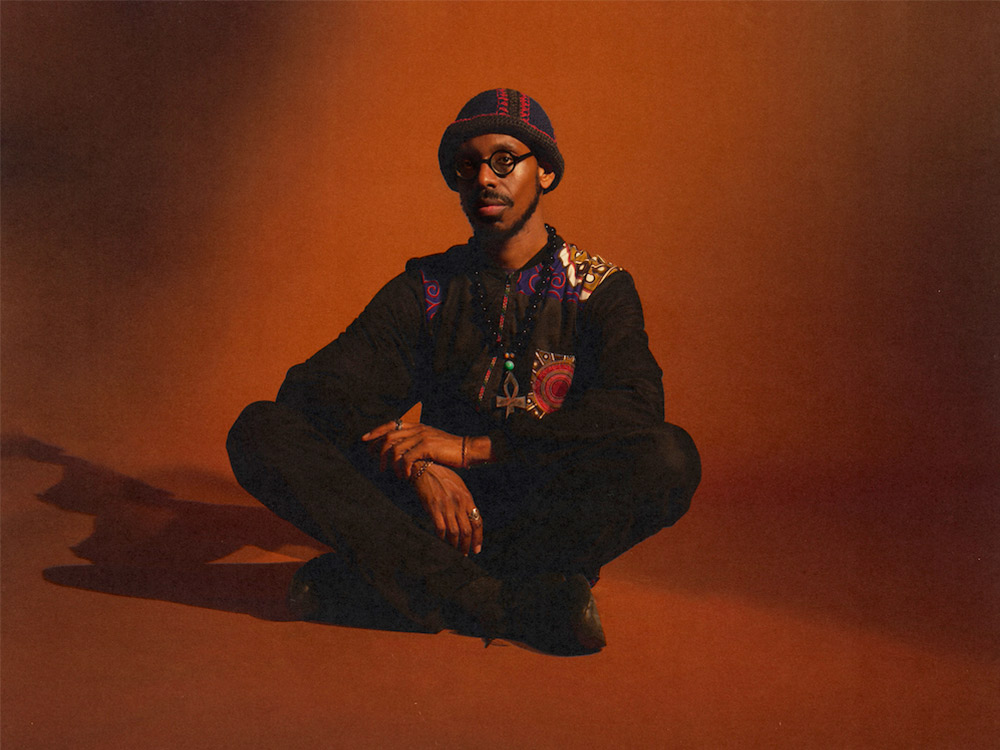 Shabaka Hutchings sits in an orange-brown room. He is wearing an ankh, and colourful knitted clothes
