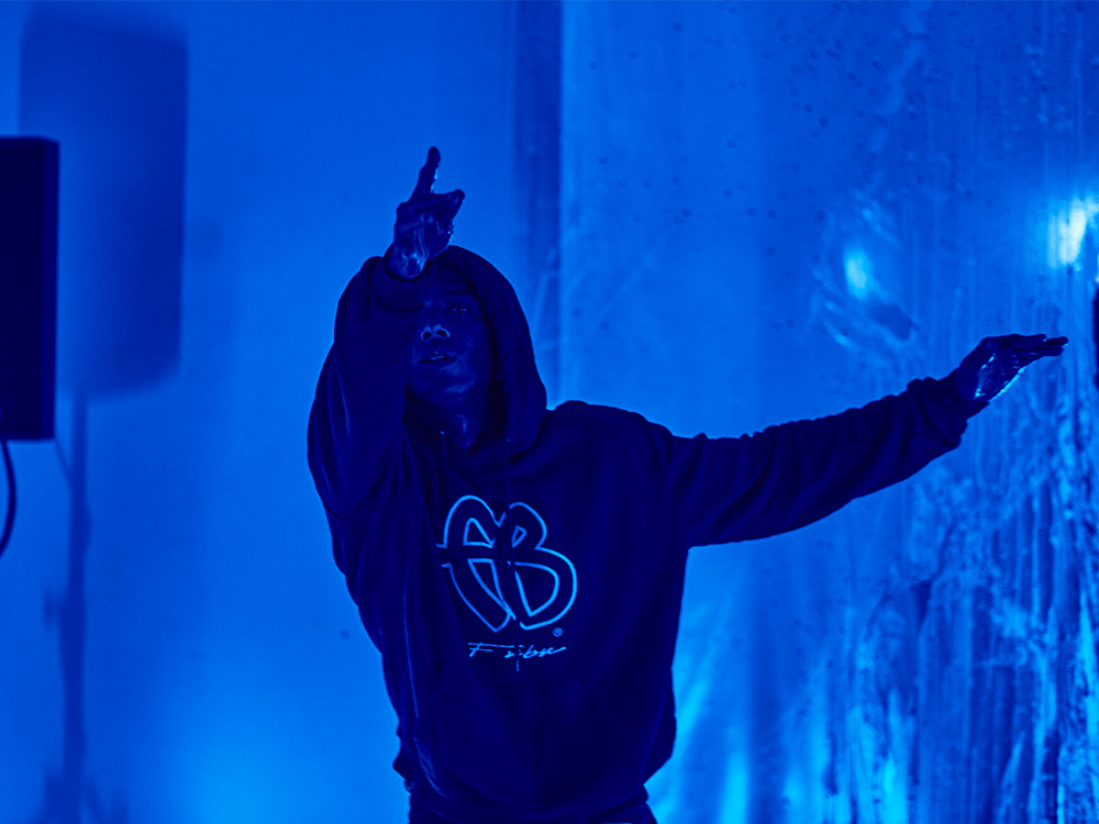A black dancer wearing a hoodie, holding their arms out against a blue background