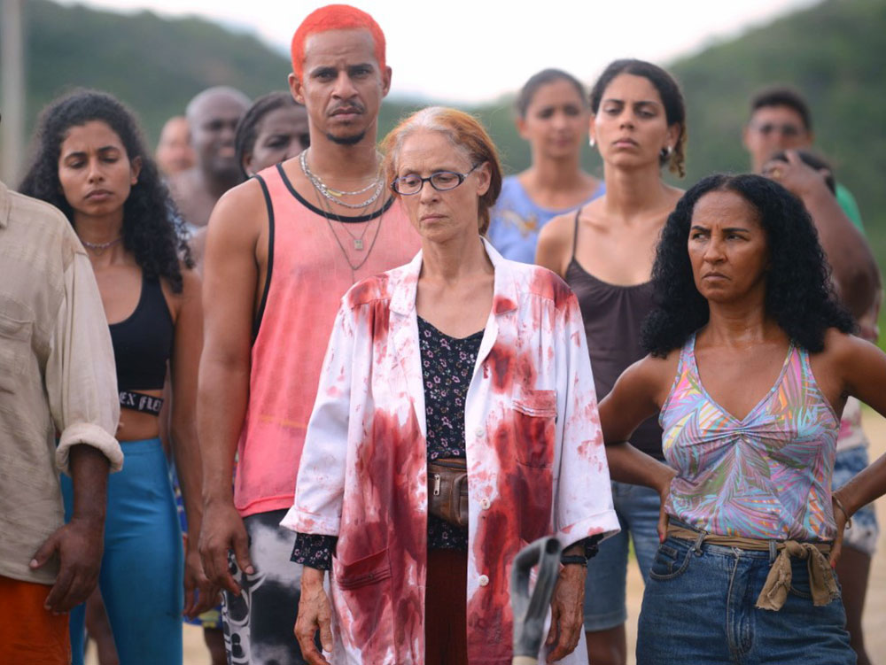 The many citizens of Bacurau stare into a hole in the ground, pictured offscreen. The older person at the front has blood covering their white coat