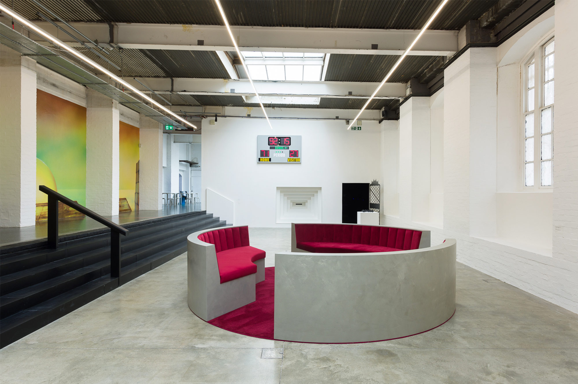 The main gallery for The Red Sun Is High, The Blue Low. On the left wall, painted stills of sunsets. Black stairs lead down to the gallery floor, where three sofas sit in a circle, with plush red velvet covering and floor. On the back wall is a baseball-like scoreboard above a black doorway and a peephole