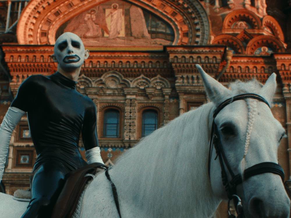 A person in latext and white face paint rides a white horse in front of a religious Russian building