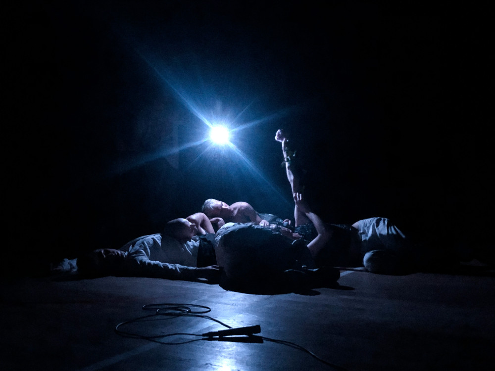 A group of people lie down under a single white light, performing Mica Levi's star star star