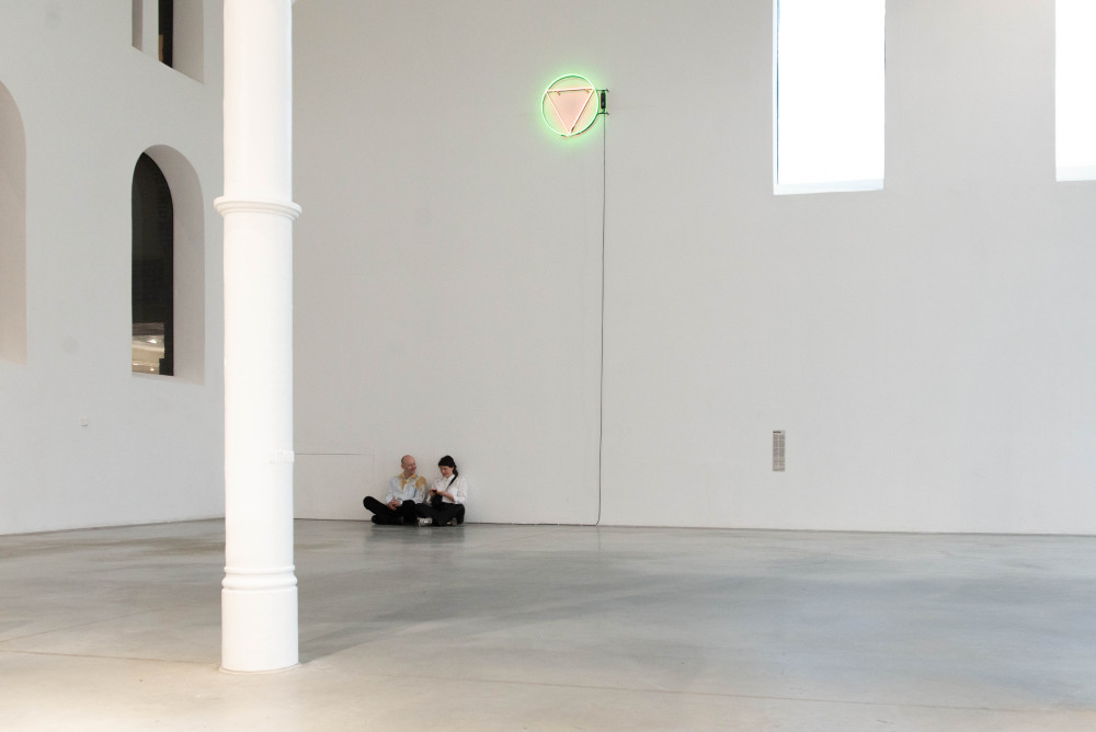 Two people sit in a sparse white gallery with arch windows. Above them is a high, green neon circle with an upside-down red triangle in it